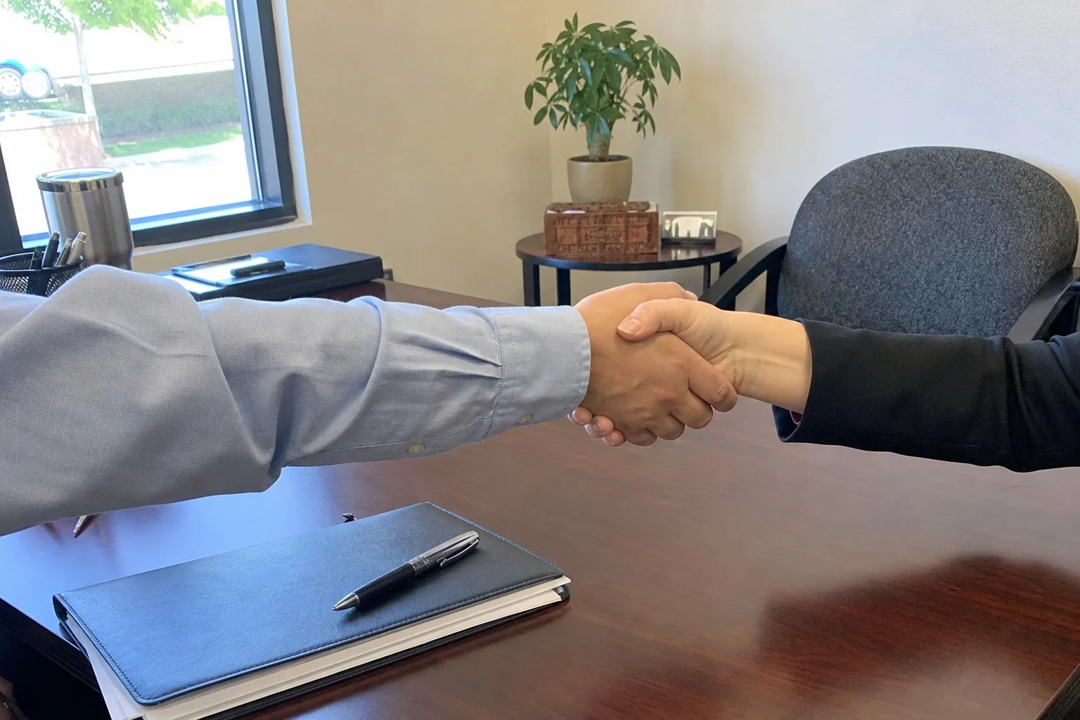 Two people shaking hands across a wooden office desk with a dark portfolio folder and pen, a window, potted plant and gray office chair behind