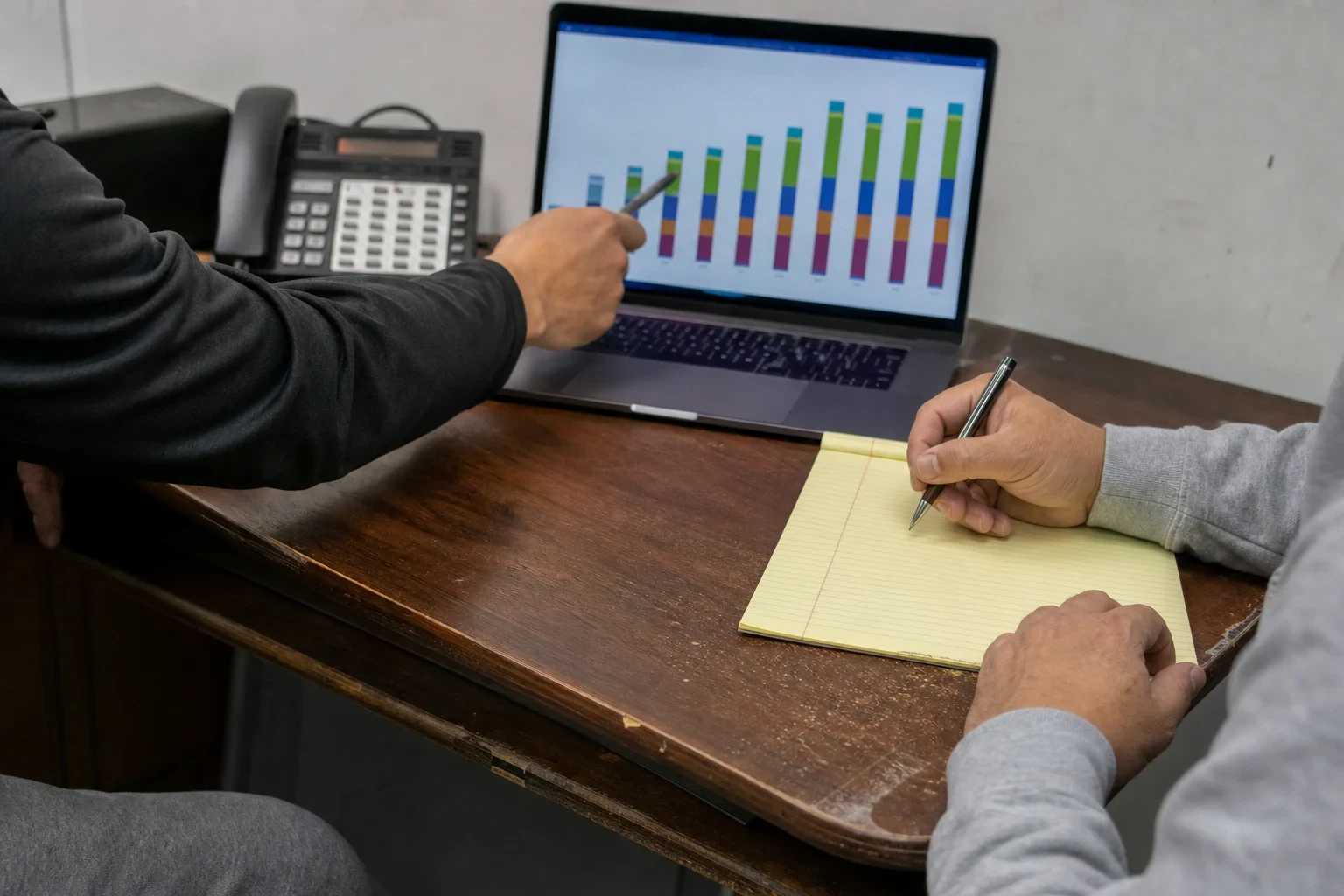 One person points a pen at a bar chart on a laptop while another takes notes on a yellow legal pad at a worn wooden office desk beside a desk phone.