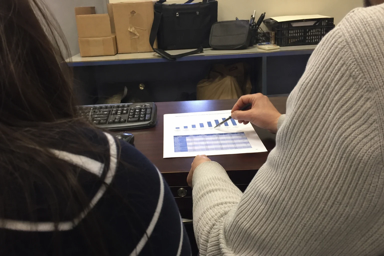 Over-the-shoulder view of two people at a back office desk as one points a pen at a printed bar chart and table, with a keyboard, boxes and bags on a shelf behind.