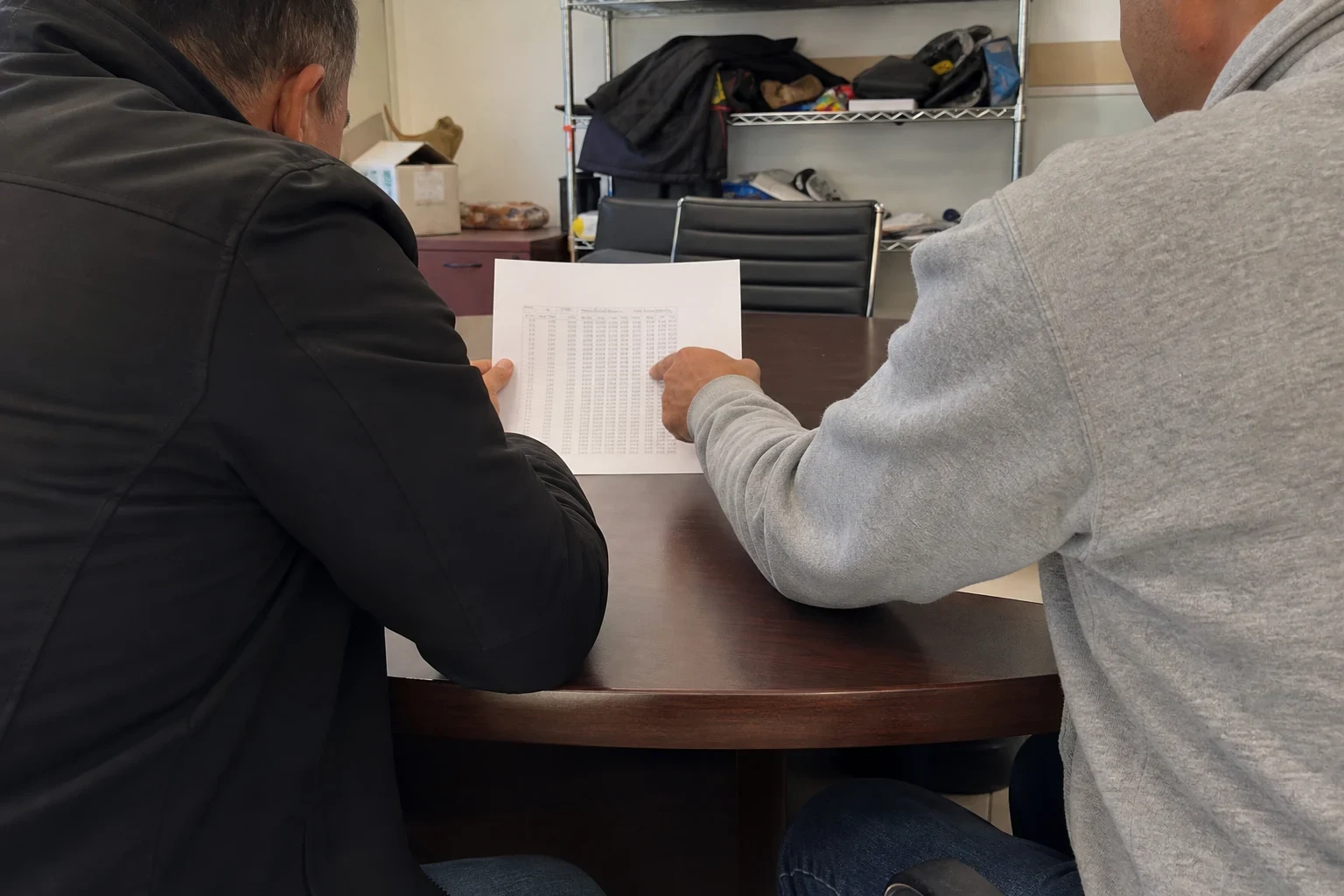 Two men seen from behind at a dark round conference table reviewing a printed page of figures, with a wire shelf and office chair in a small back office