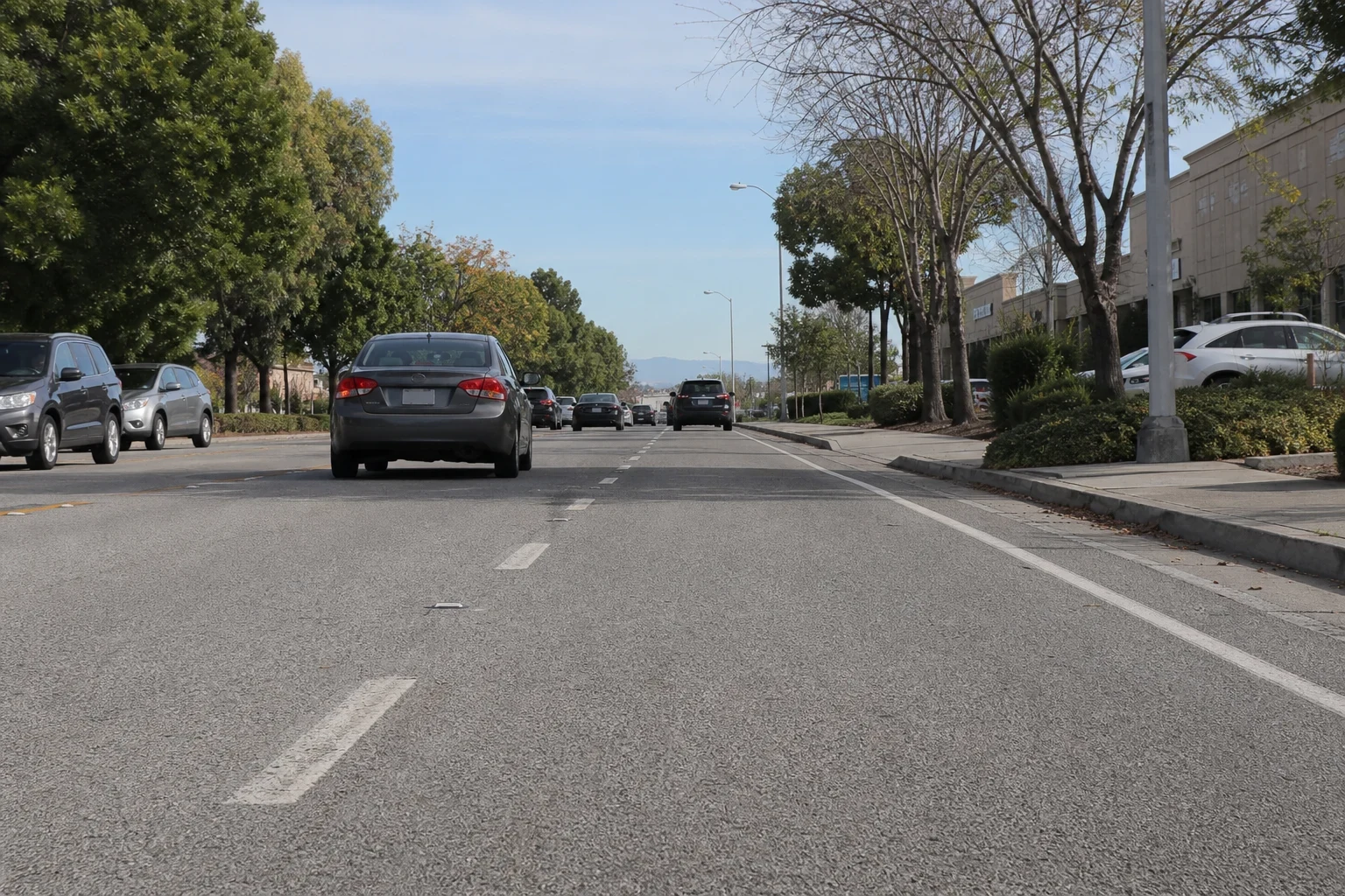 Low-angle view down a multi-lane suburban street with light traffic, trees, a sidewalk and a beige office building, with hills in the distance